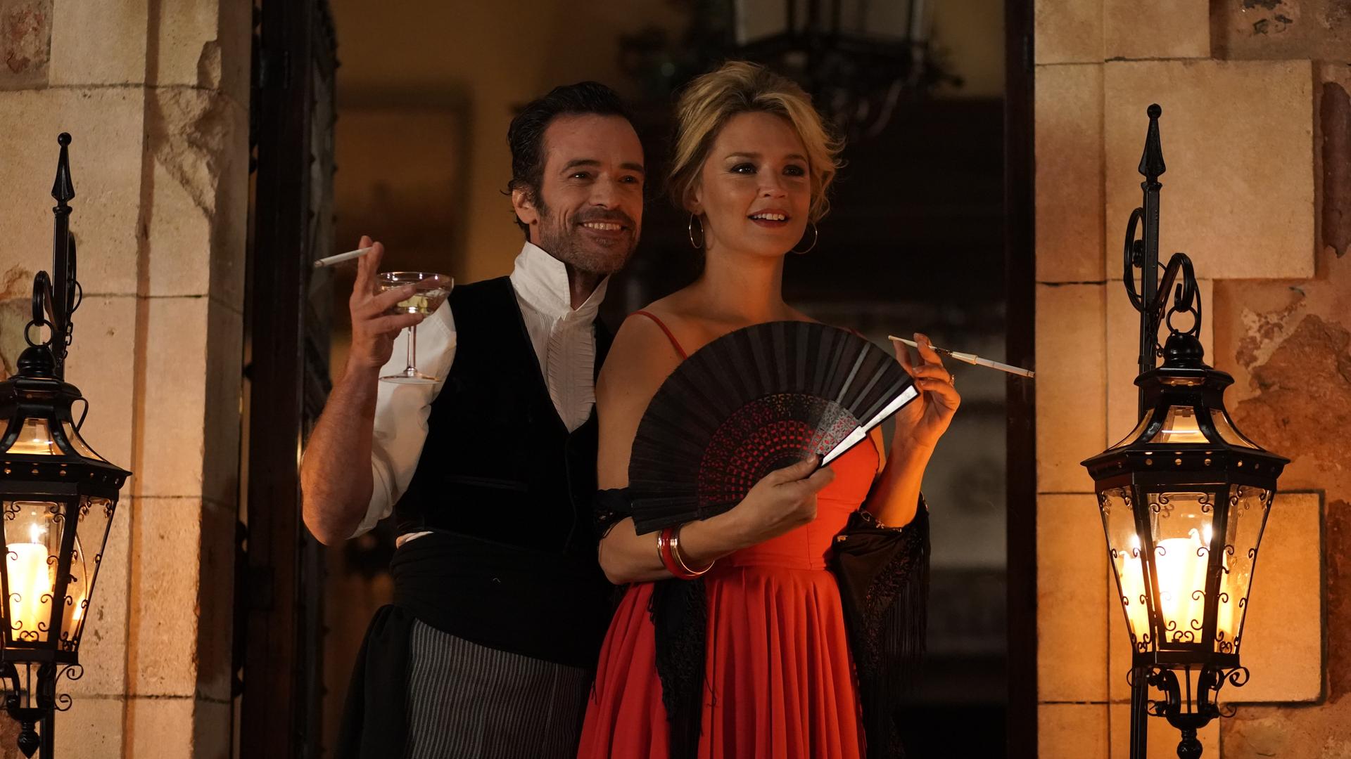 Szene aus "Warten auf Bojangles": Ein Mann im Hemd und Weste hält ein Weinglas und eine Zigarette in der rechten Hand und lacht. Links neben ihm steht eine Frau in einem roten Abendkleid. Sie hält einen Fächer. Beide stehen auf einem Balkon und schauen in die Ferne.