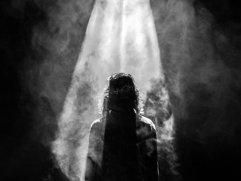 Ein ikonografisches Schwarz-Weiß-Bild einer Jesusfigur, von hinten gesehen, vor hellem Licht, das von oben auf sie herabscheint.