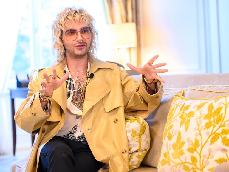 Tokio-Hotel-Sänger Bill Kaulitz sitzt in einer Suite des Hotel Vier Jahreszeiten in Hamburg auf einem Sessel und schaut in die Kamera. Er hat blonde, lockige Haare, trägt eine Brille und einen gelblichen Mantel, das Hemd ist weit offen.