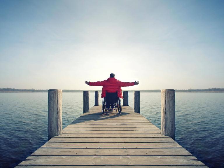 Ein Rollstuhlfahrer auf einem Bootssteg an einem See breitet die Arme aus.