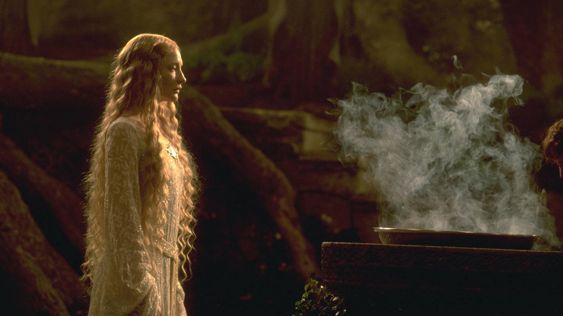 Cate Blanchett als Galadriel steht in einem weißen Kleid vor einem rauchenden Kessel. Cate Blanchett als Galadriel steht in einem weißen Kleid vor einem rauchenden Kessel.