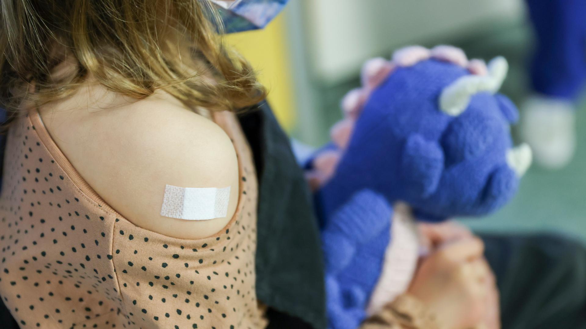 Ein Pflaster klebt auf den Arm eines Mädchens, das gegen  Covid-19 geimpft wurde. Das Kind hält einen blauen Stoffdrachen im Arm.