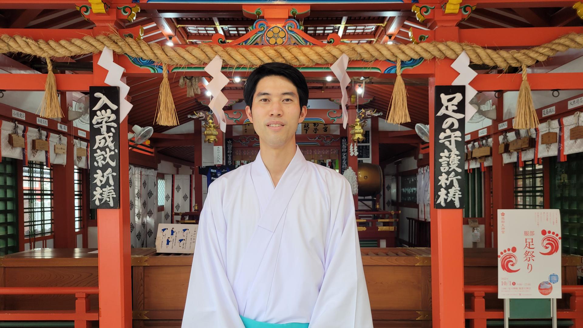 Der japanische Shinto-Priester Taishi Kato trägt ein weites weißes Hemd und einen hellblauen Rock, er steht vor dem Hattori Tenjin-gu Schrein in Osaka, einem rot gestrichenen Holzbau. Der japanische Shinto-Priester Taishi Kato trägt ein weites weißes Hemd und einen hellblauen Rock, er steht vor dem Hattori Tenjin-gu Schrein in Osaka, einem rot gestrichenen Holzbau.