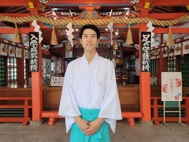Der japanische Shinto-Priester Taishi Kato trägt ein weites weißes Hemd und einen hellblauen Rock, er steht vor dem Hattori Tenjin-gu Schrein in Osaka, einem rot gestrichenen Holzbau.