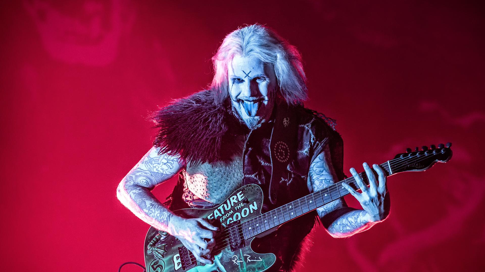Guitarist John 5 steht auf einer feuerrot ausgeleuchteten Bühne mit weiß geschminkten Gesicht wie Armen und schwarzen Markierungen auf der Stirn und streckt beim Spiel seiner Gitarre, die wild beklebt ist, die Zunge heraus und lacht dabei diabolisch. Guitarist John 5 steht auf einer feuerrot ausgeleuchteten Bühne mit weiß geschminkten Gesicht wie Armen und schwarzen Markierungen auf der Stirn und streckt beim Spiel seiner Gitarre, die wild beklebt ist, die Zunge heraus und lacht dabei diabolisch.