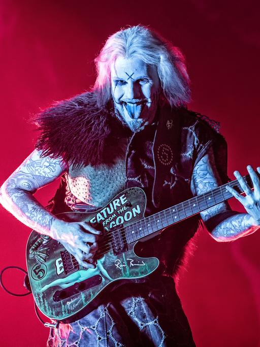 Guitarist John 5 steht auf einer feuerrot ausgeleuchteten Bühne mit weiß geschminkten Gesicht wie Armen und schwarzen Markierungen auf der Stirn und streckt beim Spiel seiner Gitarre, die wild beklebt ist, die Zunge heraus und lacht dabei diabolisch.