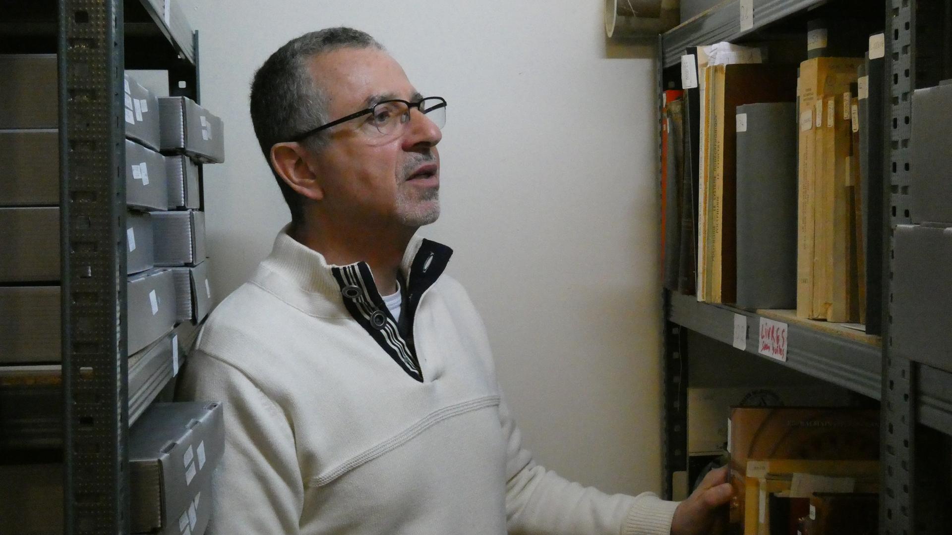 Ein Mann mit kurzem, ergrauten Haar und Brille vor einem Archiv-Regal Ein Mann mit kurzem, ergrauten Haar und Brille vor einem Archiv-Regal