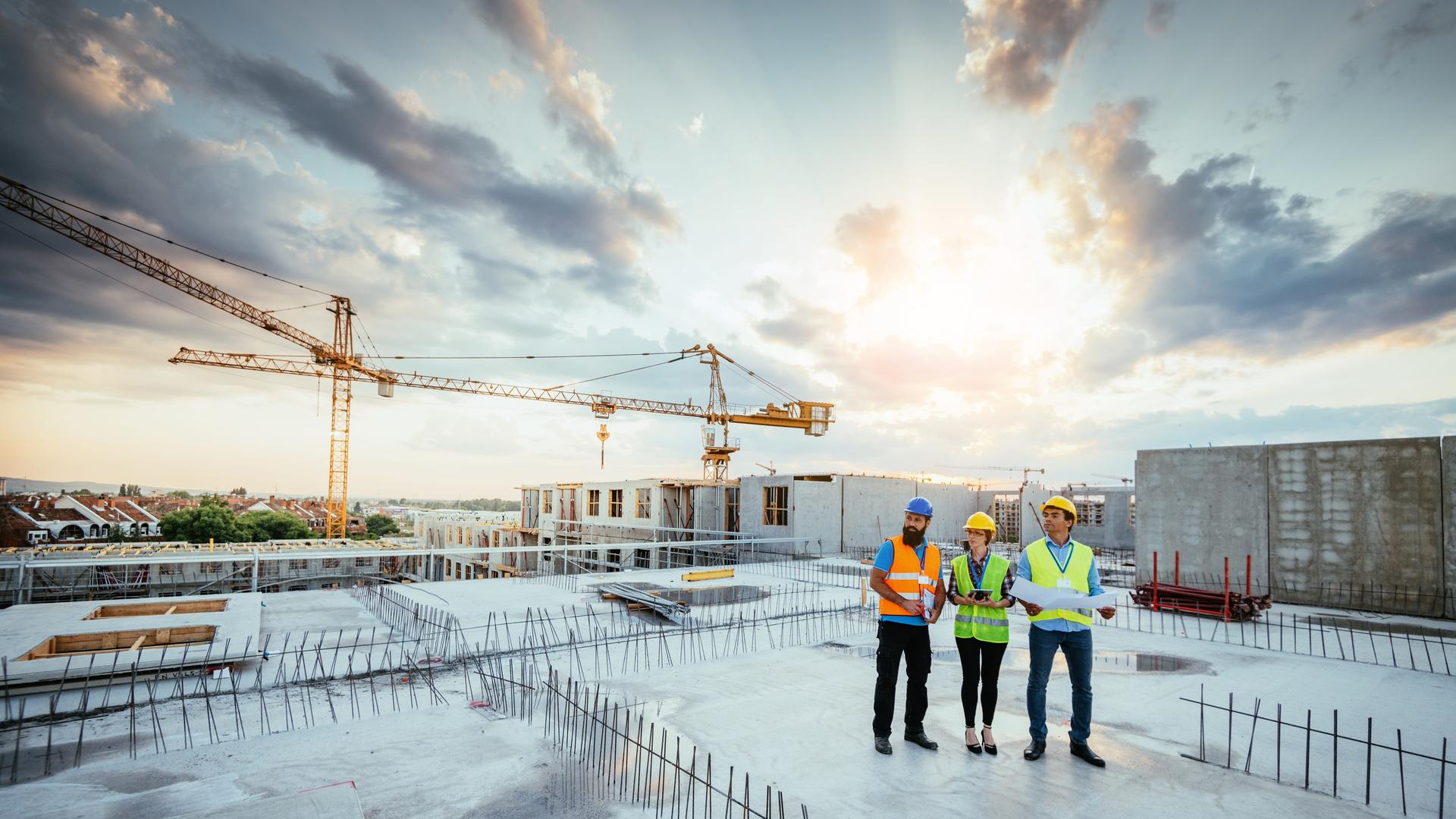Drei Personen in Warnwesten stehen auf dem Dach einer Baustelle. Hinter ihnen geht die Sonne auf. Drei Personen in Warnwesten stehen auf dem Dach einer Baustelle. Hinter ihnen geht die Sonne auf.
