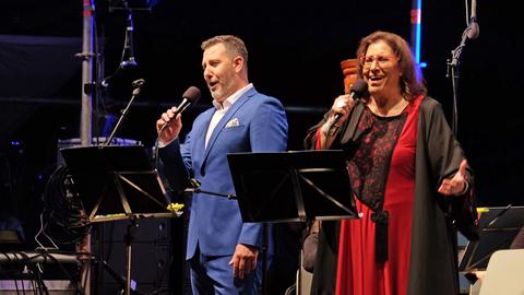 Ein Mann im blauen Anzug und eine Frau in einem schwarz-roten Kleid stehen auf einer Bühne, vor einem Orchester und singen. Sie halten Mikrofone in der Hand.