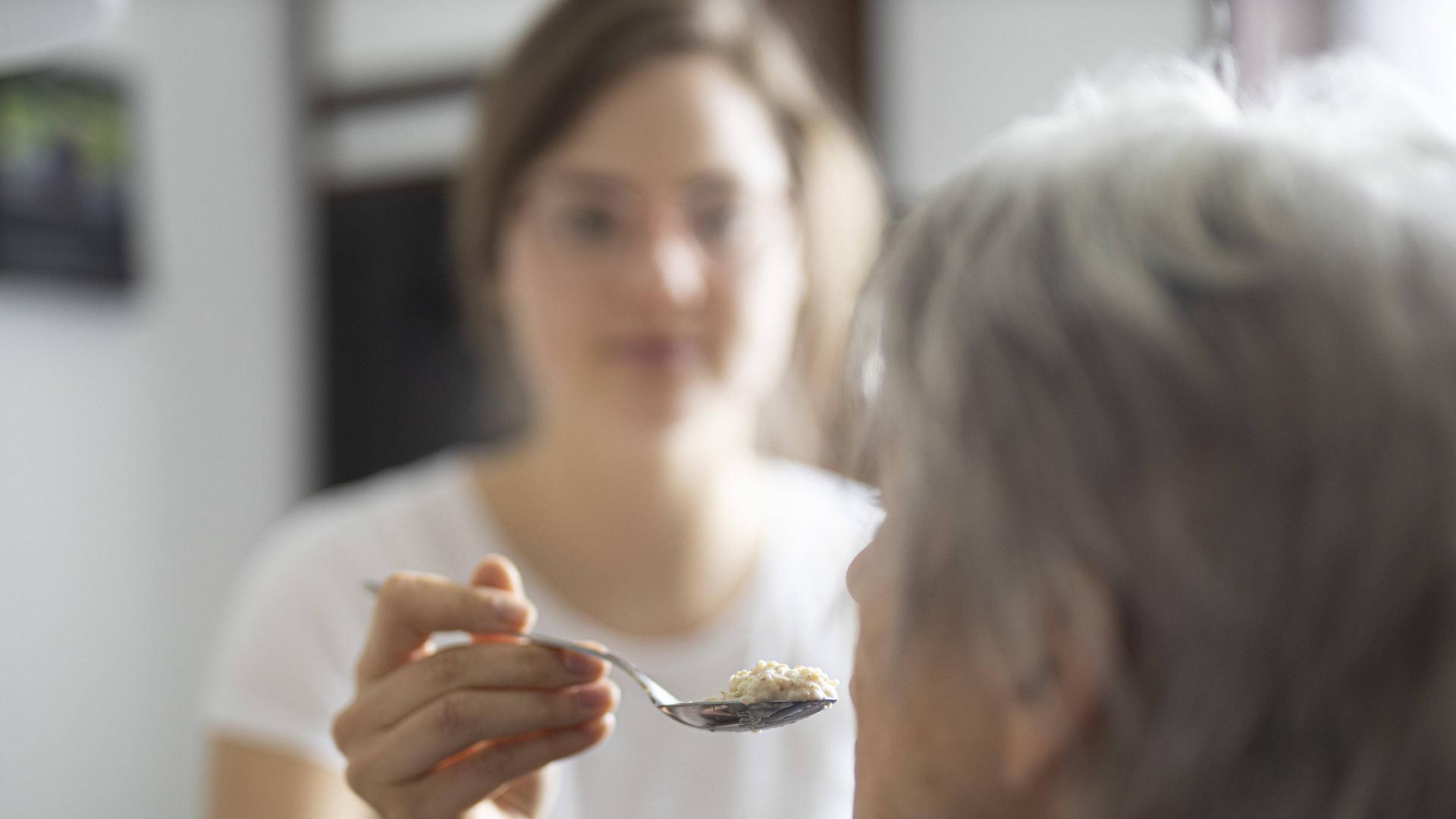 Eine alte Frau wird gefuettert. Ihr Kopf ist im Halbprofil von hinten zu sehen, sie hat graue Haare. Unscharf dahinter ist eine jüngere Frau zu sehen, die der älteren Frau einen Löffel zum Mund führt. Eine alte Frau wird gefuettert. Ihr Kopf ist im Halbprofil von hinten zu sehen, sie hat graue Haare. Unscharf dahinter ist eine jüngere Frau zu sehen, die der älteren Frau einen Löffel zum Mund führt.