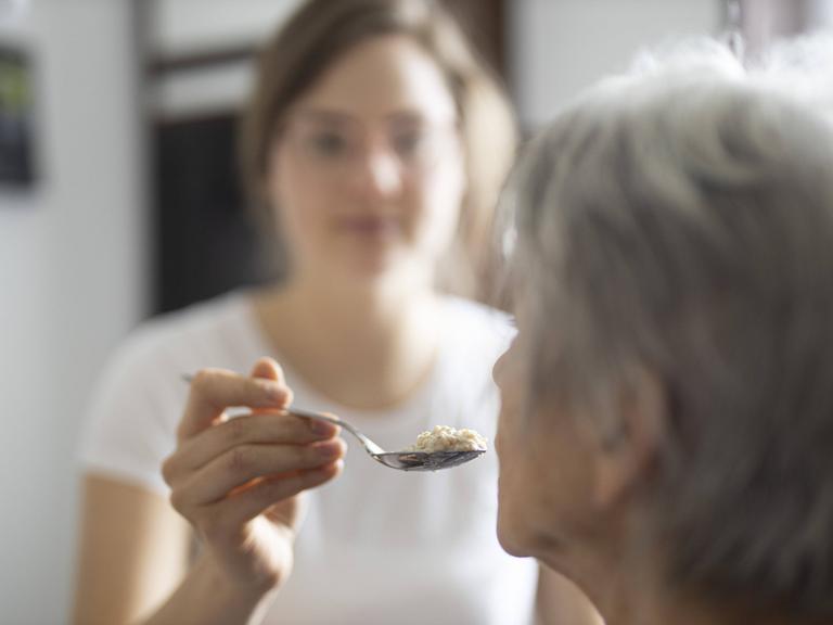 Eine alte Frau wird gefuettert. Ihr Kopf ist im Halbprofil von hinten zu sehen, sie hat graue Haare. Unscharf dahinter ist eine jüngere Frau zu sehen, die der älteren Frau einen Löffel zum Mund führt.