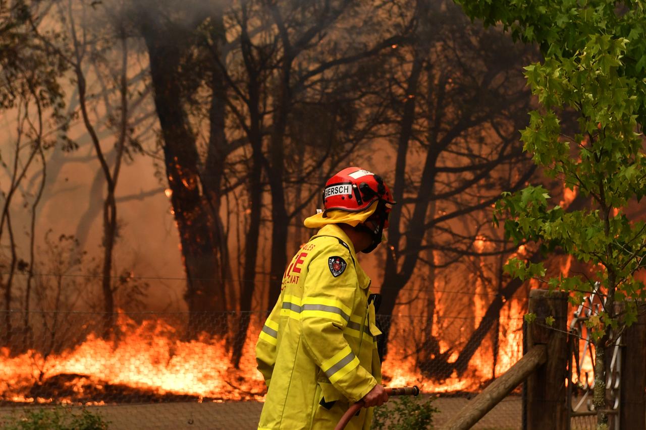 Ein Mann in gelber Jacke und mit Feuerwehrschlauch steht vor einem brennenden Stück Wald.