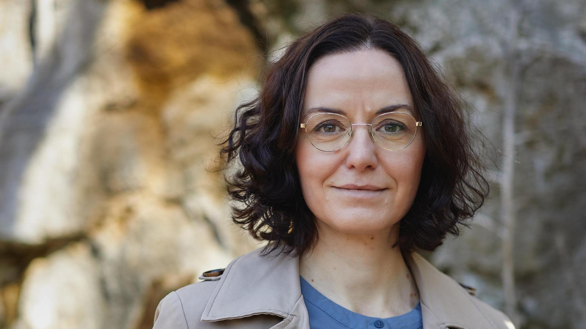 Porträt der Schriftstellerin Tina Pruschmann. Eine Frau mittleren Alters mit dunklen, halblangen Locken, goldener Brille steht im Trenchcoat und blauer Bluse vor einen teils sonnenbeschienenen Steinwand. Porträt der Schriftstellerin Tina Pruschmann. Eine Frau mittleren Alters mit dunklen, halblangen Locken, goldener Brille steht im Trenchcoat und blauer Bluse vor einen teils sonnenbeschienenen Steinwand.
