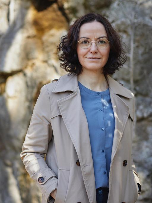 Porträt der Schriftstellerin Tina Pruschmann. Eine Frau mittleren Alters mit dunklen, halblangen Locken, goldener Brille steht im Trenchcoat und blauer Bluse vor einen teils sonnenbeschienenen Steinwand.