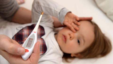 Ein digitales Fieberthermometer zeigt 37,6 Grad Celsius, dahinter ist ein Kleinkind zu sehen.