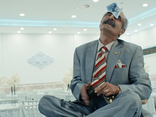 Ein türkischer Musiker im Anzug sitzt auf einem Tisch mit einem Geldschein auf der Stirn und einem Musikinstrument im Arm.
