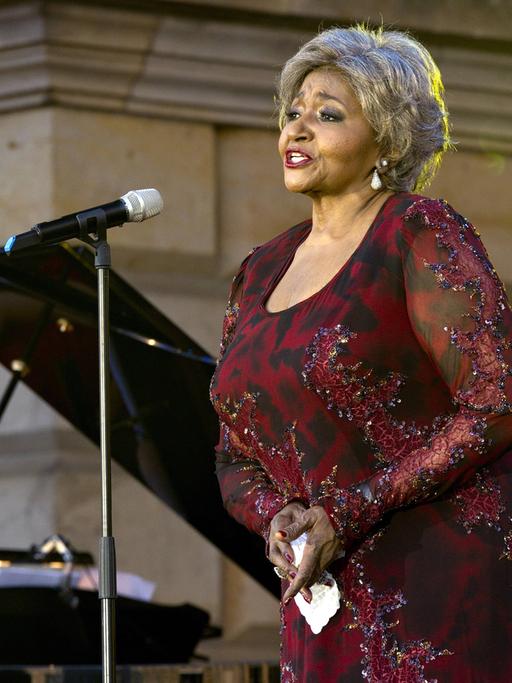 Die Opernsängerin Grace Bumbry steht in einem dunkelroten Abendkleid auf der Bühne vor einem geöffneten Flügel und singt.
