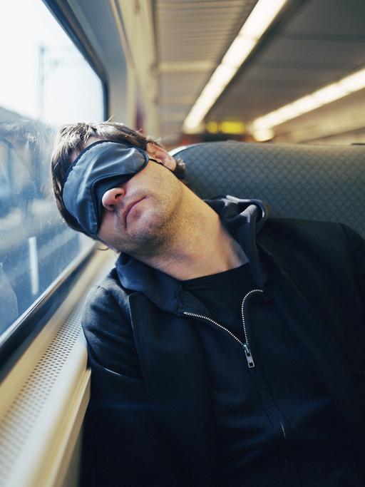 Ein junger Mann sitzt schlafend im Zug mit einer Maske vor den Augen.