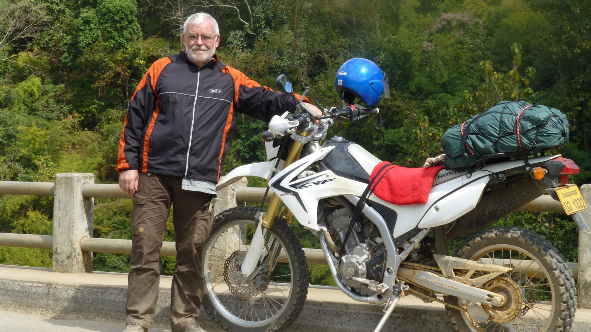 Friedrich Weidner steht neben seinem voll bepackten Motorrad auf einer Straße. Friedrich Weidner steht neben seinem voll bepackten Motorrad auf einer Straße.