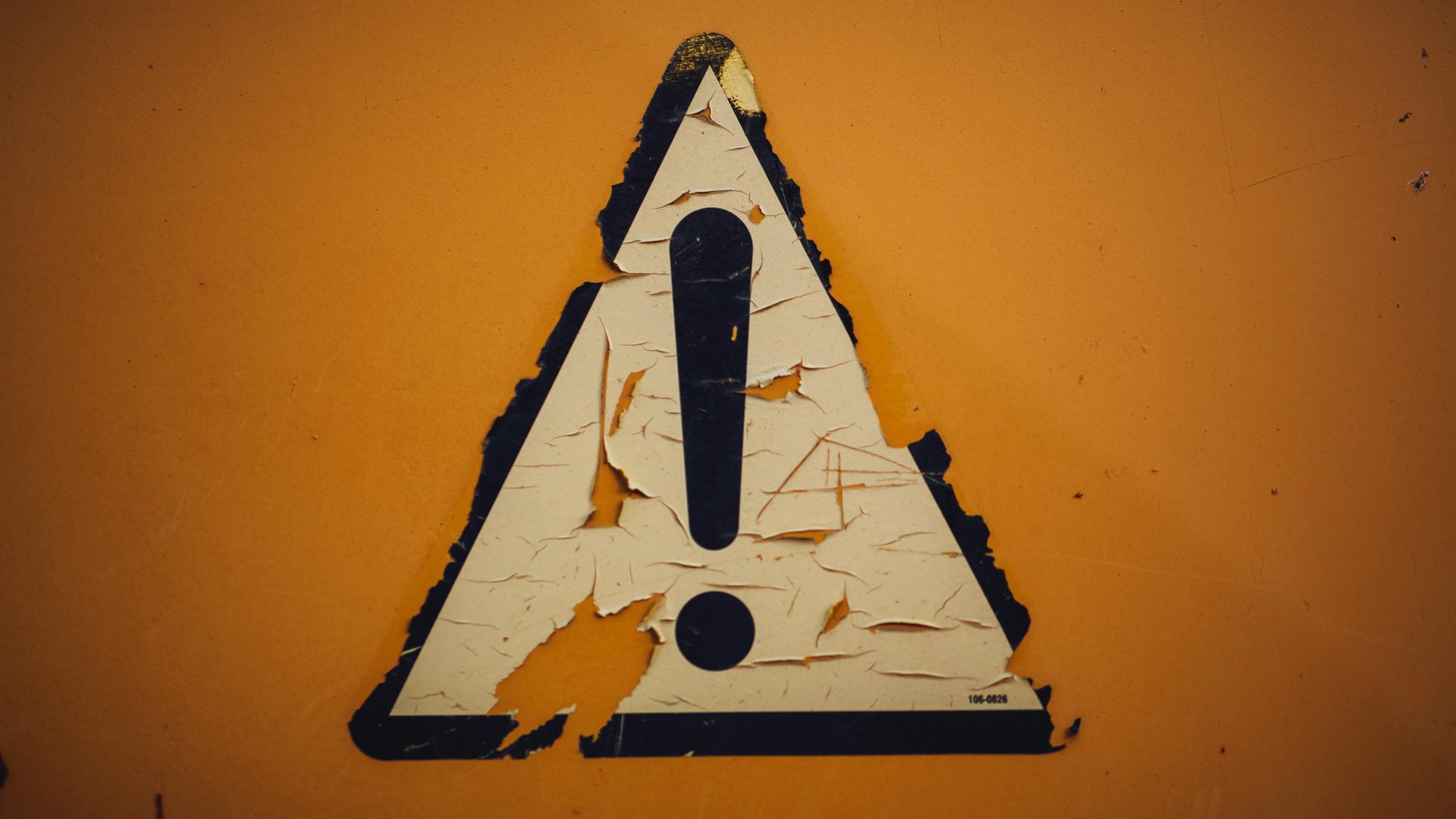 Auf gelbem Hintergrund klebt ein schwarz umrandetes weißes dreieckiges Schild mit einem Ausrufezeichen. Auf gelbem Hintergrund klebt ein schwarz umrandetes weißes dreieckiges Schild mit einem Ausrufezeichen.