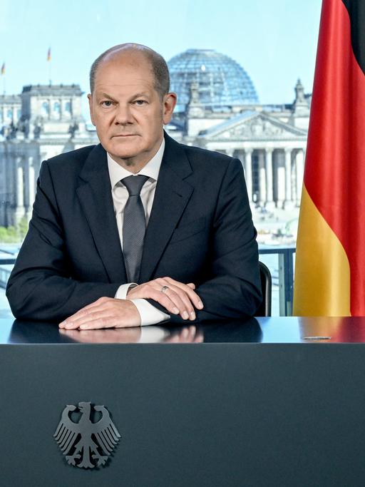 Kanzler Scholz sitzt hinter einem Pult mit Bundesadler. Neben ihm die deutsche Fahne unb die Europaflagge, im Hintergrund ist der Reichstag zu sehen. 