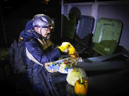 Der Jesuit Jörg Alt ist begleitet mit einer Outdoor-Jacke, er träg auch einen Helm und eine Stirnlampe. Aus einer Mülltonne holt er Lebensmittel.
