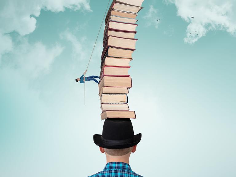 Ein Mann läuft an einem Seil einen Bücherstapel hoch, der bei einem anderen Mann auf dem Kopf steht.
