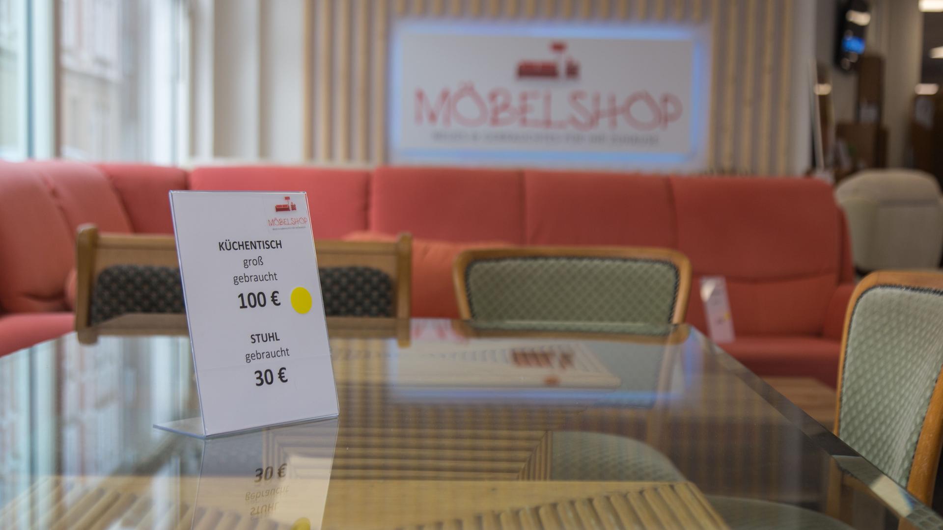 Ein Preisschild auf einem Tisch mit Glasplatte. Auf dem Schild steht: Küchentisch groß, gebraucht, 100 Euro, Stuhl, gebraucht, 30 Euro. Im Hintergrund ist unscharf die Aufschrift "Möbelshop" zu erkennen.