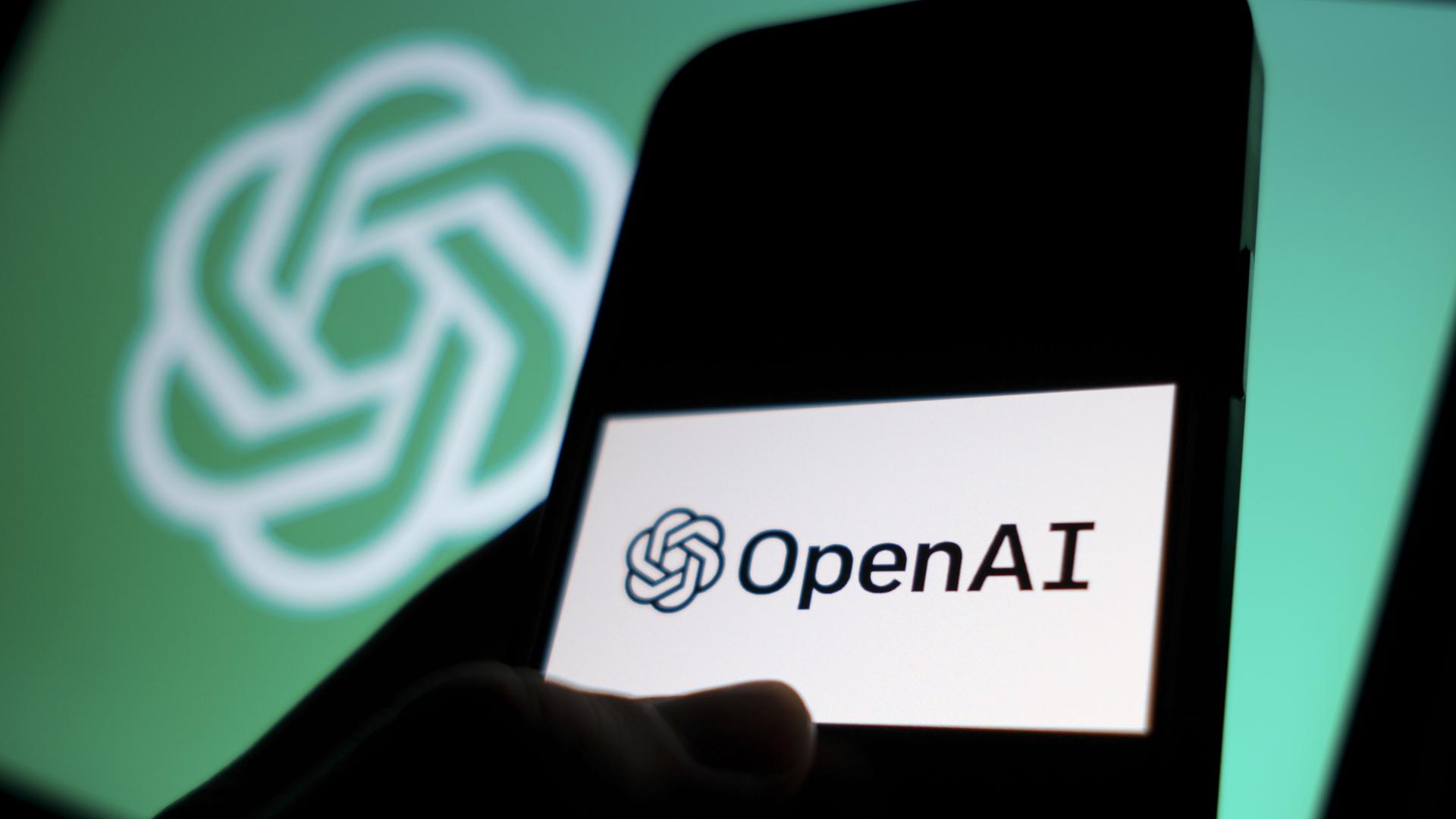 DAs Logo von"OpenAI" auf einem Smartphone, dahinter das Logo der Texterzeugungs-Ki auf einem Computer-Bildschirm