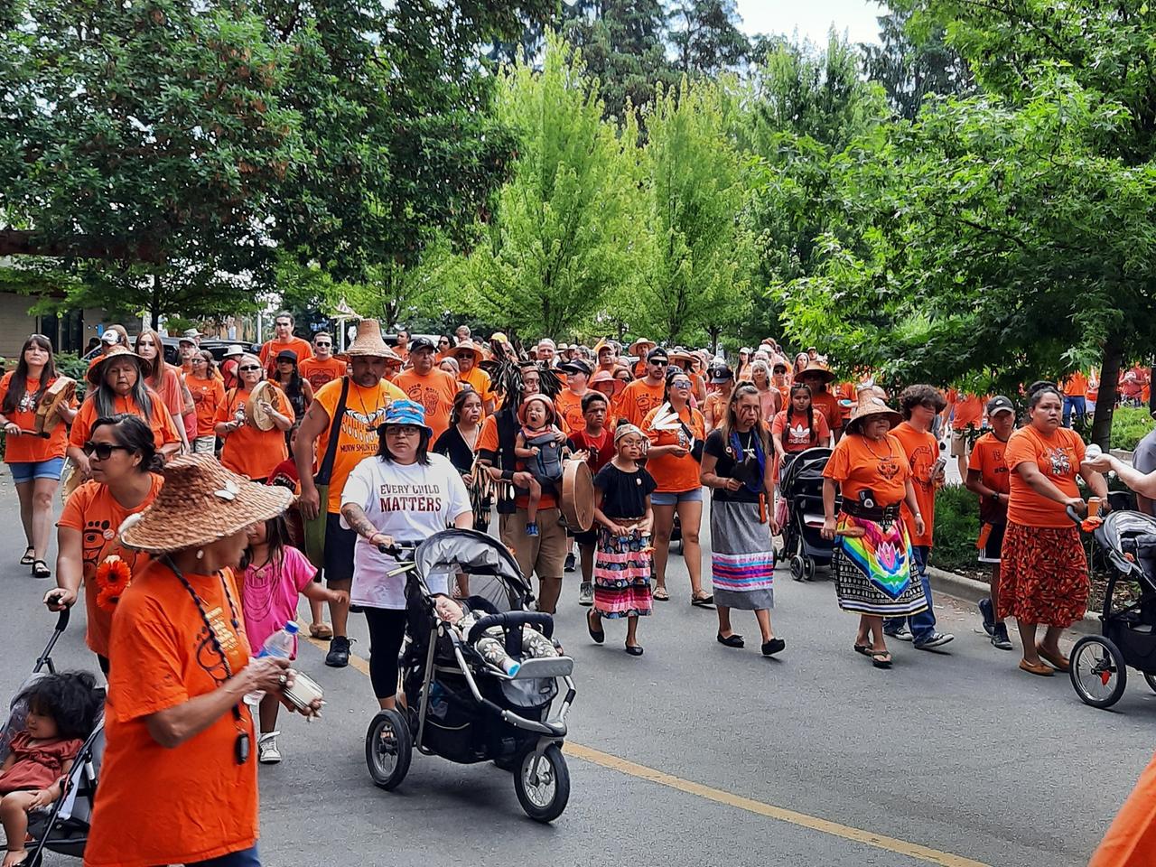 Männer, Frauen und Kinder laufen protestierend durch die Straßen. Sie tragen orangene T-Shirts, zum Teil mit der Aufschrift "Every Child Matters". 