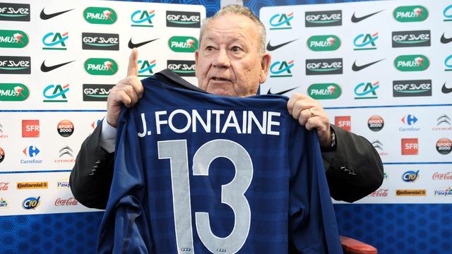 Das Foto zeigt den ehemaligen Fußball-Spieler Just Fontaine aus dem Land Frankreich. Er hält ein blaues Trikot mit dem Namen Fontaine und der Nummer 13 hoch.