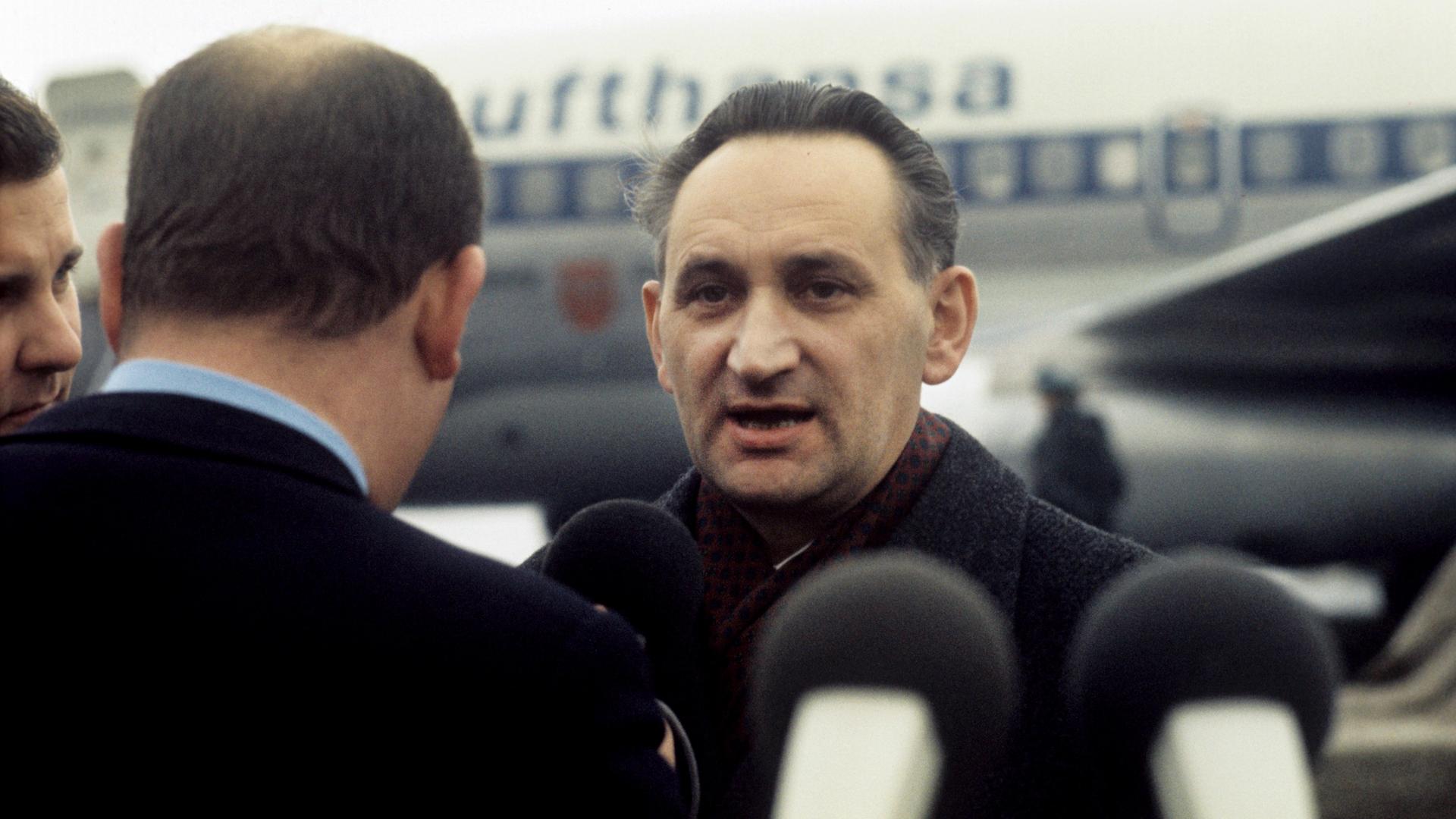 Ein Mann steht umringt von Reportern auf einem Flugfeld. Hinter ihm: ein Flugzeug. Ein Mann steht umringt von Reportern auf einem Flugfeld. Hinter ihm: ein Flugzeug.