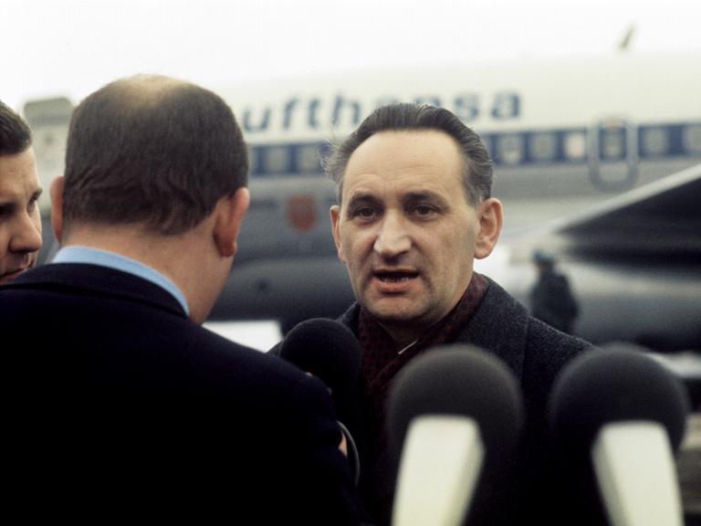 Ein Mann steht umringt von Reportern auf einem Flugfeld. Hinter ihm: ein Flugzeug.