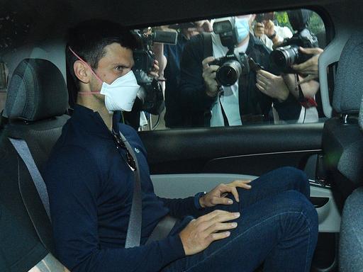 Ein Mann mit Mund-Nasen-Schutz sitzt auf dem Rücksitz eines Autos. Am Wagenfenster stehen Fotografen und fotografieren ihn.