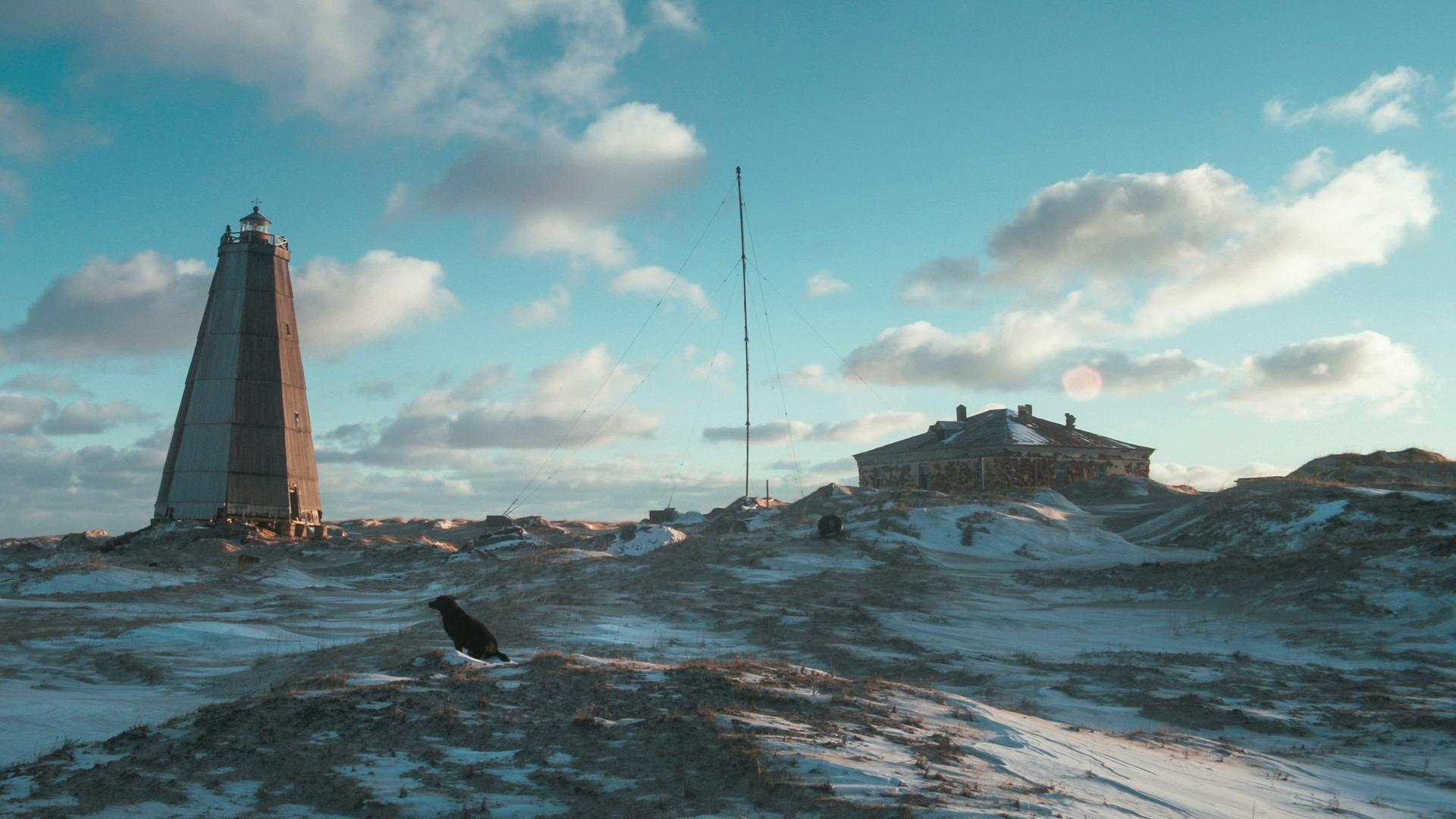 In einer verschneiten Lanschaft steht ein alter Leuchtturm im Morgenlicht, auf einem Hügel im Vordergrund sitzt ein schwarzer Hund. In einer verschneiten Lanschaft steht ein alter Leuchtturm im Morgenlicht, auf einem Hügel im Vordergrund sitzt ein schwarzer Hund.
