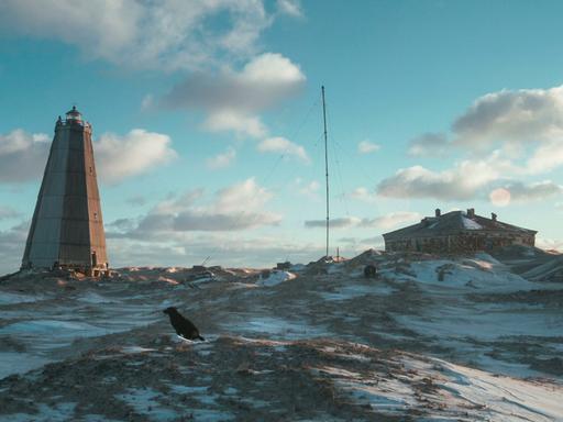 In einer verschneiten Lanschaft steht ein alter Leuchtturm im Morgenlicht, auf einem Hügel im Vordergrund sitzt ein schwarzer Hund.