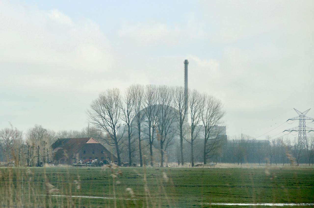 Eine leicht dunst-nebelige Landschaft mit Feld und Bäumen. In der Ferne ist schemenhaft die typische Rundkuppel eines Atomkraftwerkes zu erkennen, daneben ein hoher Schornstein.
