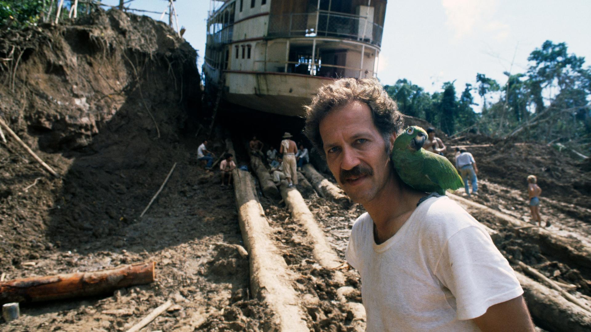 Werner Herzog am Set zu "Fitzcarraldo" mit einem grünen Papageien auf der Schulter und einem auf Baumstämmen transportierten Dampfschiff im Hintergrund. Werner Herzog am Set zu "Fitzcarraldo" mit einem grünen Papageien auf der Schulter und einem auf Baumstämmen transportierten Dampfschiff im Hintergrund.