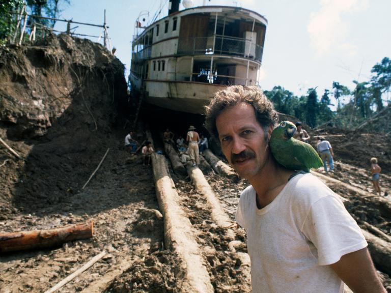 Werner Herzog am Set zu "Fitzcarraldo" mit einem grünen Papageien auf der Schulter und einem auf Baumstämmen transportierten Dampfschiff im Hintergrund.
