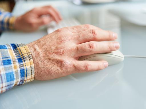 Eine Hand im Vordergrund bedient eine Computermaus. Die Hand im Hintergrund liegt auf der Computer-Tastatur. 