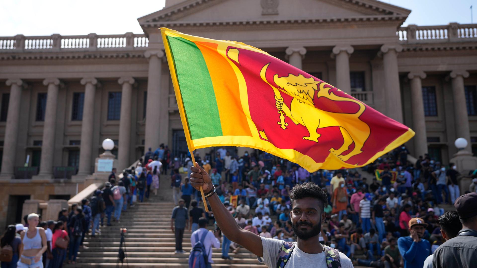 Menschen stehen vor dem Präsidenten-Palast in Colombo in Sri Lanka. Im Vordergrund ein Mann, der eine Fahne schwenkt. Menschen stehen vor dem Präsidenten-Palast in Colombo in Sri Lanka. Im Vordergrund ein Mann, der eine Fahne schwenkt.