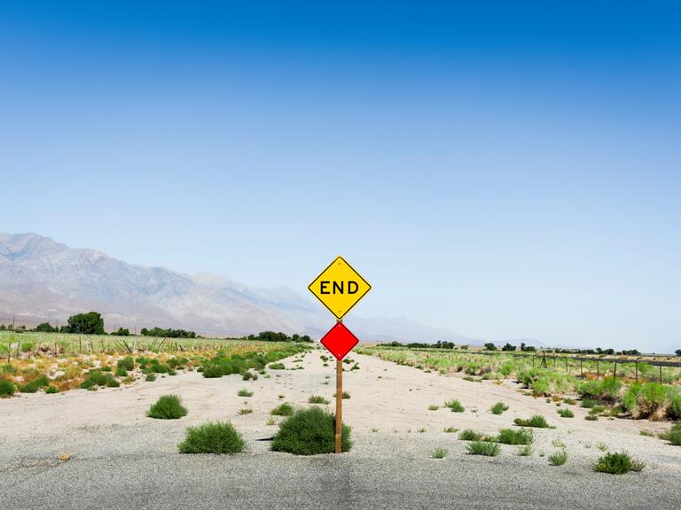 Ein Schotterweg endet in einer dürren Landschaft. Das Ende des Weges ist mit einem gelben Schild markiert, das die Aufschrift "End" trägt.