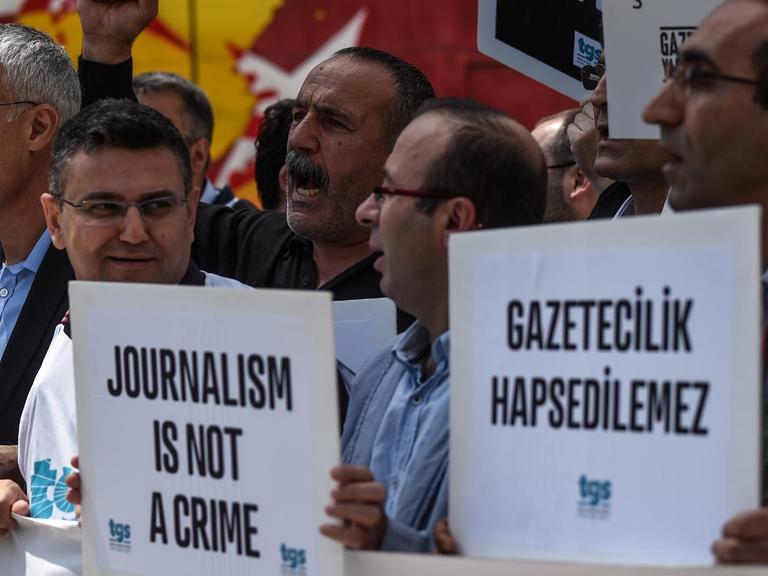 Mehrere Männer stehen in einer Menge, einige halten Plakate mit der Aufschrift "Journalismus ist kein Verbrechen".