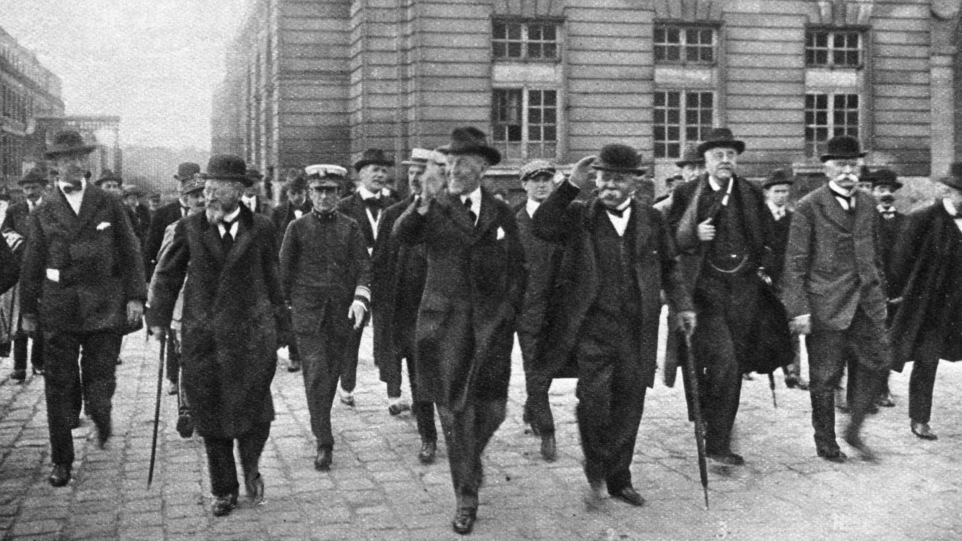 Schwarzweiß Kulisse vor der mehrere Männer in schwarzen Anzügen und Hüten laufen.