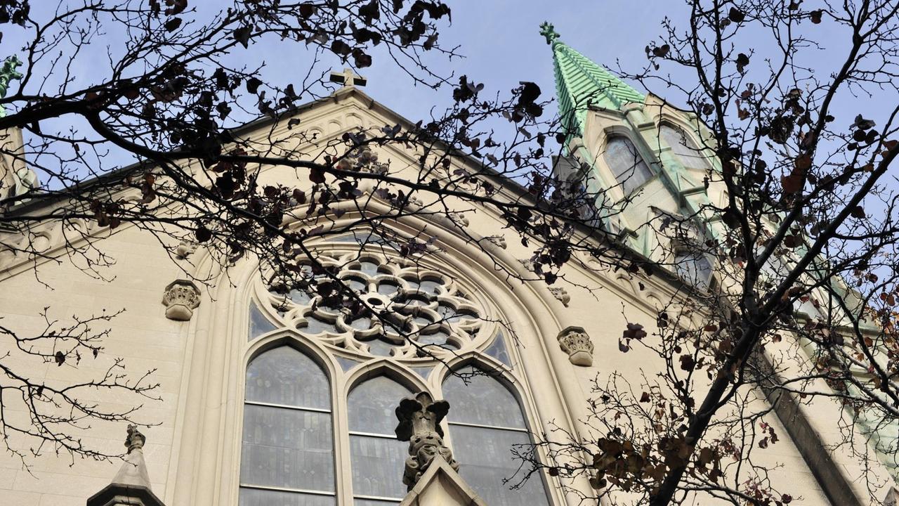 Blick durch die Äste eines Baums auf eine Kirchenfassade mit gotischen Glasfenstern und einer grünen Kirchturmspitze