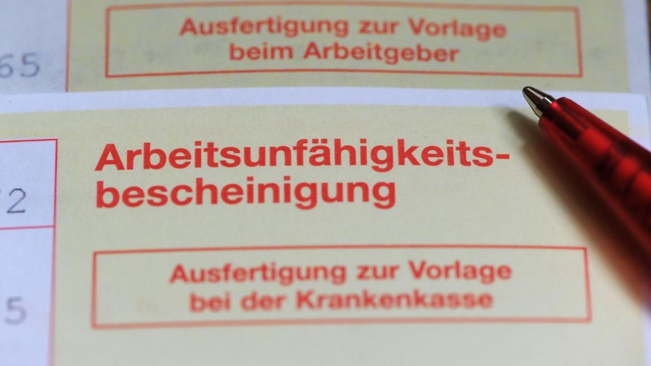 Zu sehen ist eine typisch gelb-rote Arbeitsunfähigkeitsbescheinigung, auf der ein Stift liegt. Zu sehen ist eine typisch gelb-rote Arbeitsunfähigkeitsbescheinigung, auf der ein Stift liegt.