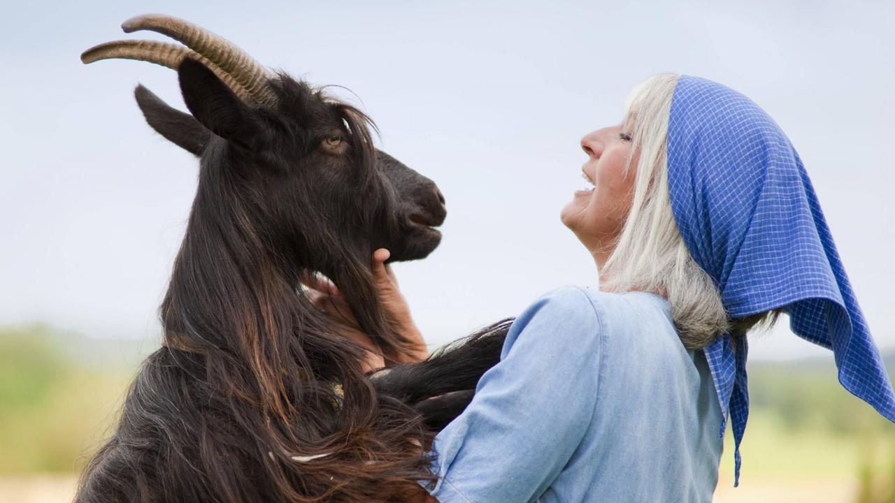 Ein Ziegenbock hat seine Vorderhufe auf die Schultern einer Frau gelegt, sie lacht ihn an. Ein Ziegenbock hat seine Vorderhufe auf die Schultern einer Frau gelegt, sie lacht ihn an.