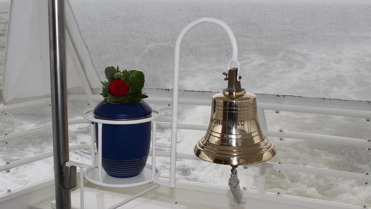 Eine Urne mit der Asche von Verstorbenen auf einem Schiff. Neben ihr hängt eine Schiffsglocke. Im Hintergrund ist die Nordsee zu sehen.