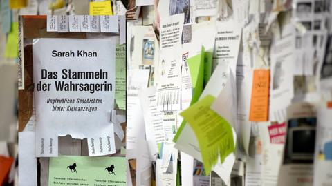 Buchcover "Das Stammeln der Wahrsagerin" von Sarah Khan vor einer Wand mit Kleinanzeigen