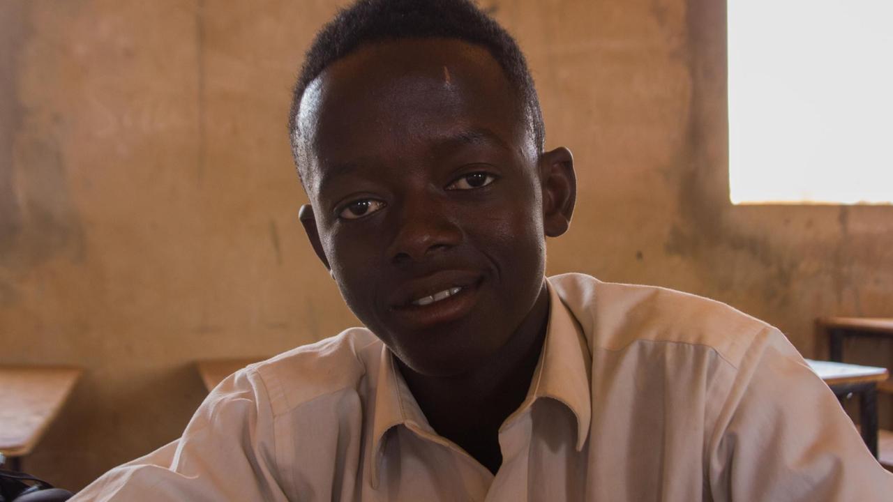 Ein afrikanischer Junge sitzt im Hemd an einem Pult im Klassenzimmer einer Schule.